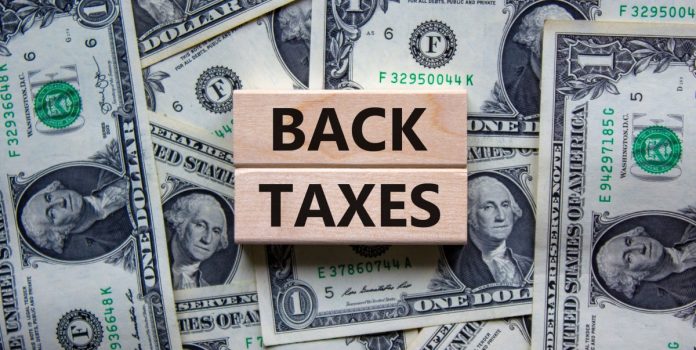 impostos-retroativos-apostas-brasil Back Taxes escrito em blocos de madeira, posicionados sobre notas de dólares
