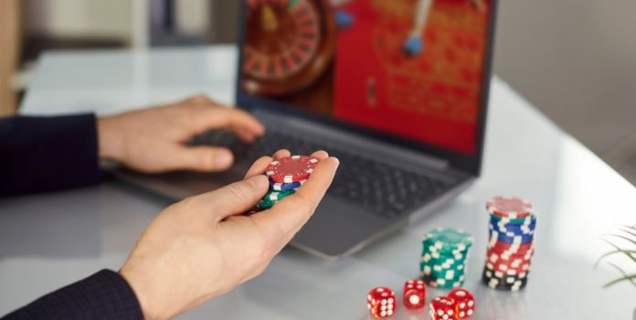 Homem sentado com fichas de cassino na mão e com computador jogando roleta