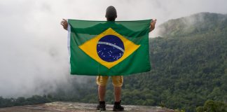 Homem na montanha segurando a bandeira do Brasil e olhando para o céu