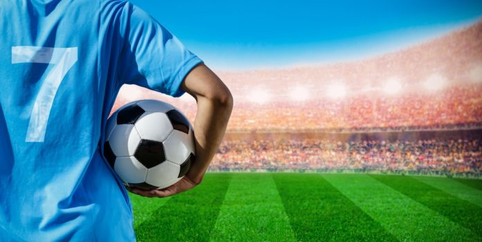 Homem segurando bola de futebol vestindo camiseta 7 em frente ao campo