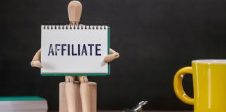 Bruna Lima, F12Bet: afiliados geram tráfego, ampliam alcance e trazem credibilidade à marca Boneco de madeira sentado segurando bloco de notas escrito affiliate