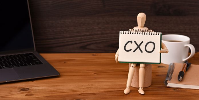 EstrelaBet-nomeia-Lorena-Lima-diretora-de-UX-e-Expansão Boneco segurando cartaz com a sigla CXO sobre a mesa, ao lado do notebook. da xícara de café e do bloco de notas