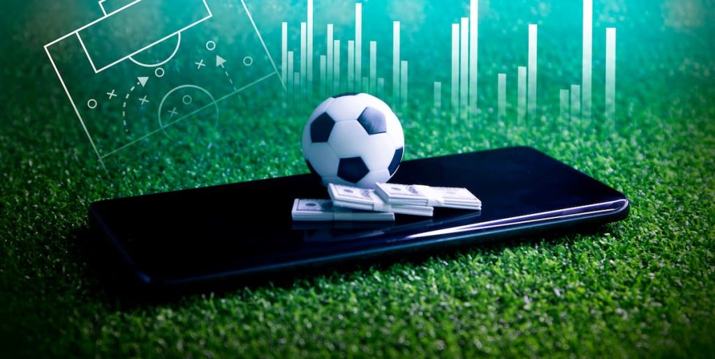 Uma bola de futebol e maços de dinheiro sob um smartphone, simbolizando apostas esportivas.