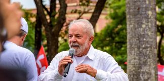 Presidente Lula, um homem branco de idade avançada e cabelos brancos, discursa com um microfone em mãos.