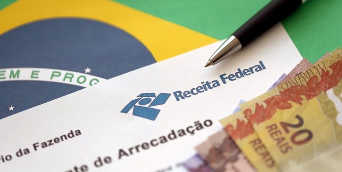 Receita-Federal Uma folha da Receita Federal com notas de 20, 10 e 5 reais apoiada na bandeira do Brasil.