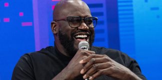 Shaquille O'Neal, homem negro de meia idade, segura um microfone enquanto dá risada.