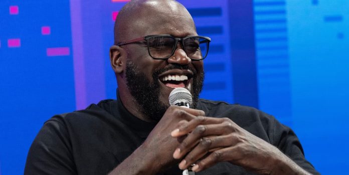 Shaquille O'Neal, homem negro de meia idade, segura um microfone enquanto dá risada.