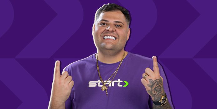 Start-Bet-Maumau-Embaixador Homem de meia-idade, vestido com uma camisa roxa escrito Start, aponta os dois dedos indicadores para cima. Trata-se do influenciador digital Maumau, novo embaixador da Start Bet.