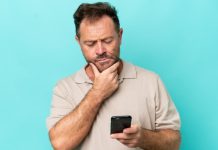 Homem mexendo no celular com uma mão, enquanto a outra está no queixo e com cara de dúvida