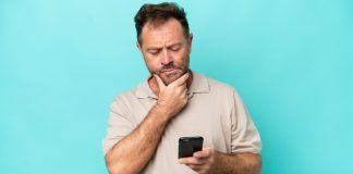 Homem mexendo no celular com uma mão, enquanto a outra está no queixo e com cara de dúvida