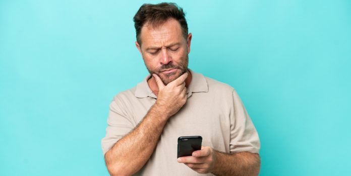 joberto-porto-espaço-jurídico-expansão-das-apostas-no-brasil Homem mexendo no celular com uma mão, enquanto a outra está no queixo e com cara de dúvida