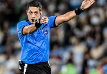 Árbitro de futebol, homem, apita e faz gesto com a mão esquerda apontando para um jogador.