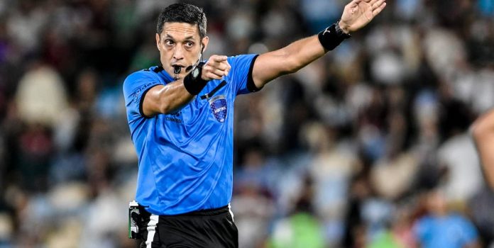 Árbitro-Futebol Árbitro de futebol, homem, apita e faz gesto com a mão esquerda apontando para um jogador.