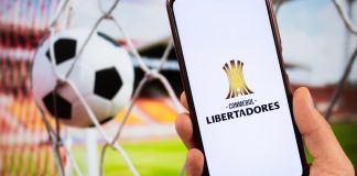 Pay4Fun: Transações financeiras em sites de aposta crescem mais de 80% próximo a jogos da Libertadores Celular com uma tela escrito Conmebol Libertadores ao lado de uma bola na rede.