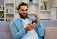 Homem barbudo, de cabelo longo, celebra enquanto segura o celular.