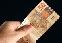 Mão masculina segurando uma nota de 50 reais com fundo preto