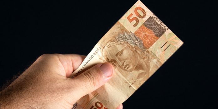 Mão masculina segurando uma nota de 50 reais com fundo preto