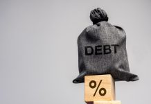 ANJL destaca contradições do Desenrola 2.0 Um saco com a escrita "debt" em cima de blocos com o símbolo da porcentagem empilhados