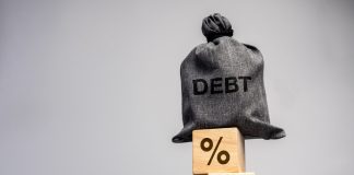 ANJL destaca contradições do Desenrola 2.0 Um saco com a escrita "debt" em cima de blocos com o símbolo da porcentagem empilhados