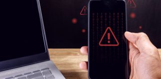 Anatel divulga dados sobre bloqueio de sites de apostas após pedido via LAI Homem segurando um smartphone com símbolo de cuidado em vermelho aparecendo na tela e um notebook desligado ao lado
