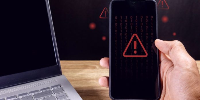 Anatel-divulga-dados-sobre-bloqueio-de-sites-de-apostas-apos-pedido-via-LAI Homem segurando um smartphone com símbolo de cuidado em vermelho aparecendo na tela e um notebook desligado ao lado