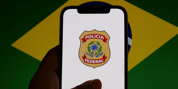 SPA-determina-repasse-de-recursos-de-apostas-ao-Funapol-via-DARF-5862 Um celular mostrando o brasão da Polícia Federal com a bandeira do Brasil desfocada ao fundo