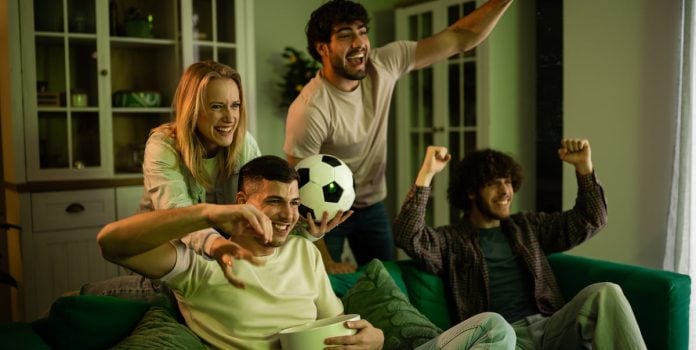 Betsul-Patrocina-Os-Donos-da-Bola-Band Grupo de torcedores assistindo TV com uma bola na mão.