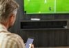 Homem assistindo futebol na TV enquanto faz apostas em seu smartphone.