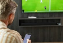 Homem assistindo futebol na TV enquanto faz apostas em seu smartphone.