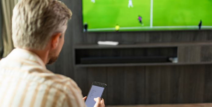 68-dos-Brasileiros-Defende-Proibicao-de-Patrocinio-de-Apostas-no-Futebol Homem assistindo futebol na TV enquanto faz apostas em seu smartphone.