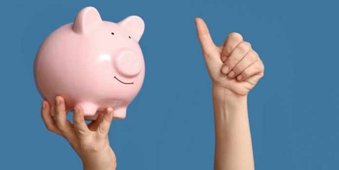 Executivos-e-especialistas-apontam-beneficios-do-setor-de-apostas-para-a-sociedade Uma pessoa segurando um cofrinho de porco com uma mão e fazendo o sinal de joinha com a outra em um fundo azul