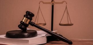 Uma mesa de tribunal com um malhete sobre um livro e a Balança da Justiça ao fundo