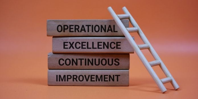 Blocos de madeira com excelência operacional e melhoria contínua escritos em inglês, formando base para escada inclinada que representa o progresso operacional