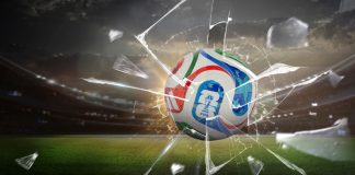 Uma imagem da bola oficial da Copa do Mundo de 2026 atravessando um vidro quebrado, com um gramado ao fundo