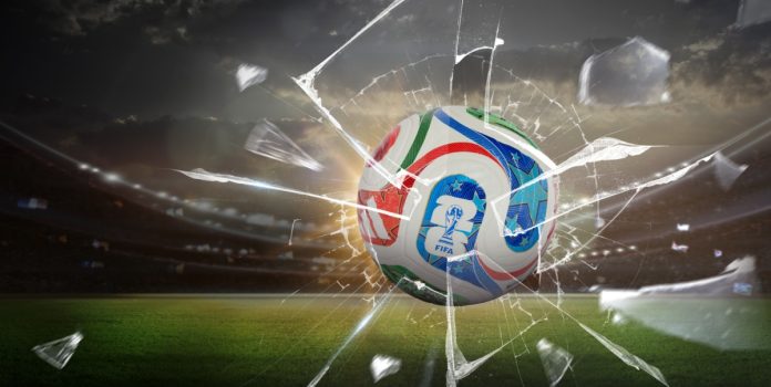 Uma imagem da bola oficial da Copa do Mundo de 2026 atravessando um vidro quebrado, com um gramado ao fundo