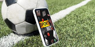 Um celular exibindo apostas esportivas apoiado em uma bola de futebol no gramado de um campo