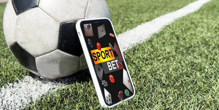 Um celular exibindo apostas esportivas apoiado em uma bola de futebol no gramado de um campo