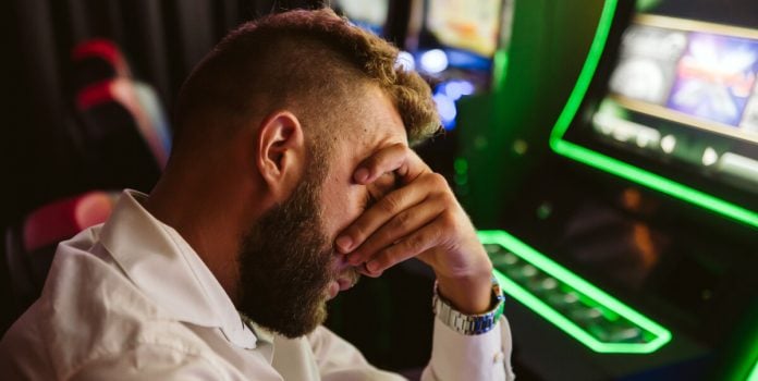 PL-Cadastro-Nacional-de-Autoexclusão-na-Lei-das-Apostas Homem triste com a mão no rosto e uma máquina de jogos no fundo
