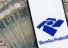 Notas de 100 reais ao lado de celular mostrando o aplicativo da Receita Federal.