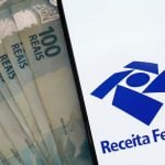 Notas de 100 reais ao lado de celular mostrando o aplicativo da Receita Federal.