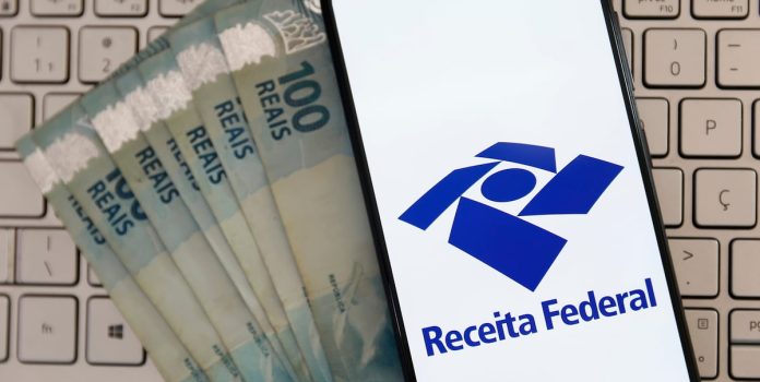 Notas de 100 reais ao lado de celular mostrando o aplicativo da Receita Federal.