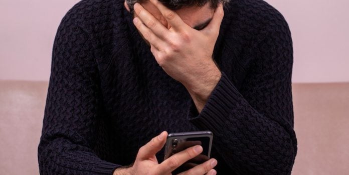 ministério-da-fazenda-divulga-dados-sobre-apostas-brasil Um homem sentado com uma mão tapando o rosto e a outra mexendo no celular