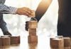 Betsul firma parceria com SOFTSWISS para ampliar portfólio Duas pessoas montando uma torre de cubos de madeira sobre uma mesa para simbolizar parceria e união