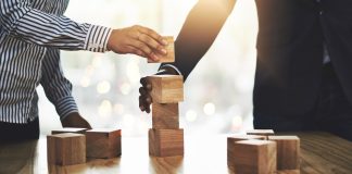 Betsul firma parceria com SOFTSWISS para ampliar portfólio Duas pessoas montando uma torre de cubos de madeira sobre uma mesa para simbolizar parceria e união