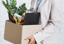 Mulher irreconhecível segurando uma caixa com pertences pessoais, saindo do escritório após abandonar o emprego.