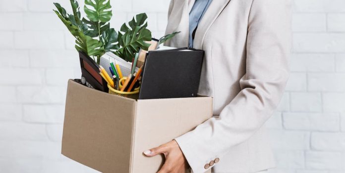 Mulher irreconhecível segurando uma caixa com pertences pessoais, saindo do escritório após abandonar o emprego.