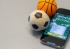 Bola de futebol e de basquete ao lado de um celular com um aplicativo de apostas esportivas.