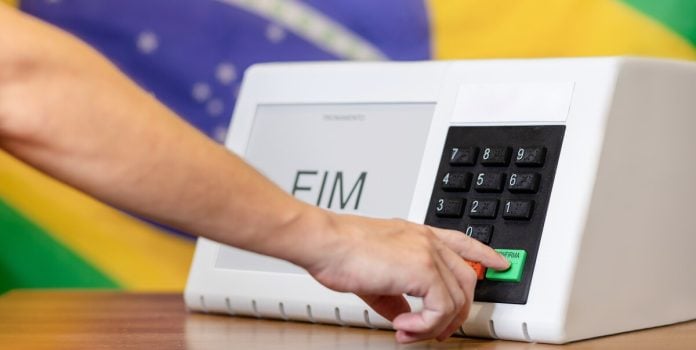 datafolha-empate-técnico-lula-flávio-bolsonaro-segundo-turno-eleições-2026 Uma pessoa votando com a bandeira do Brasil ao fundo